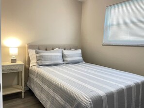 Ein Schlafzimmer mit einem Bett, einem Nachttisch, einer Lampe und einem Fenster mit Jalousien.