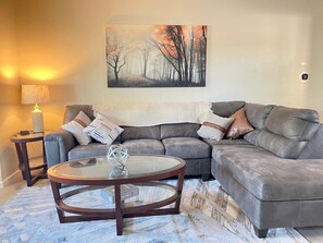 Ein Wohnzimmer mit einem Ecksofa, einem runden Couchtisch, einer Lampe und einem Gemälde an der Wand.