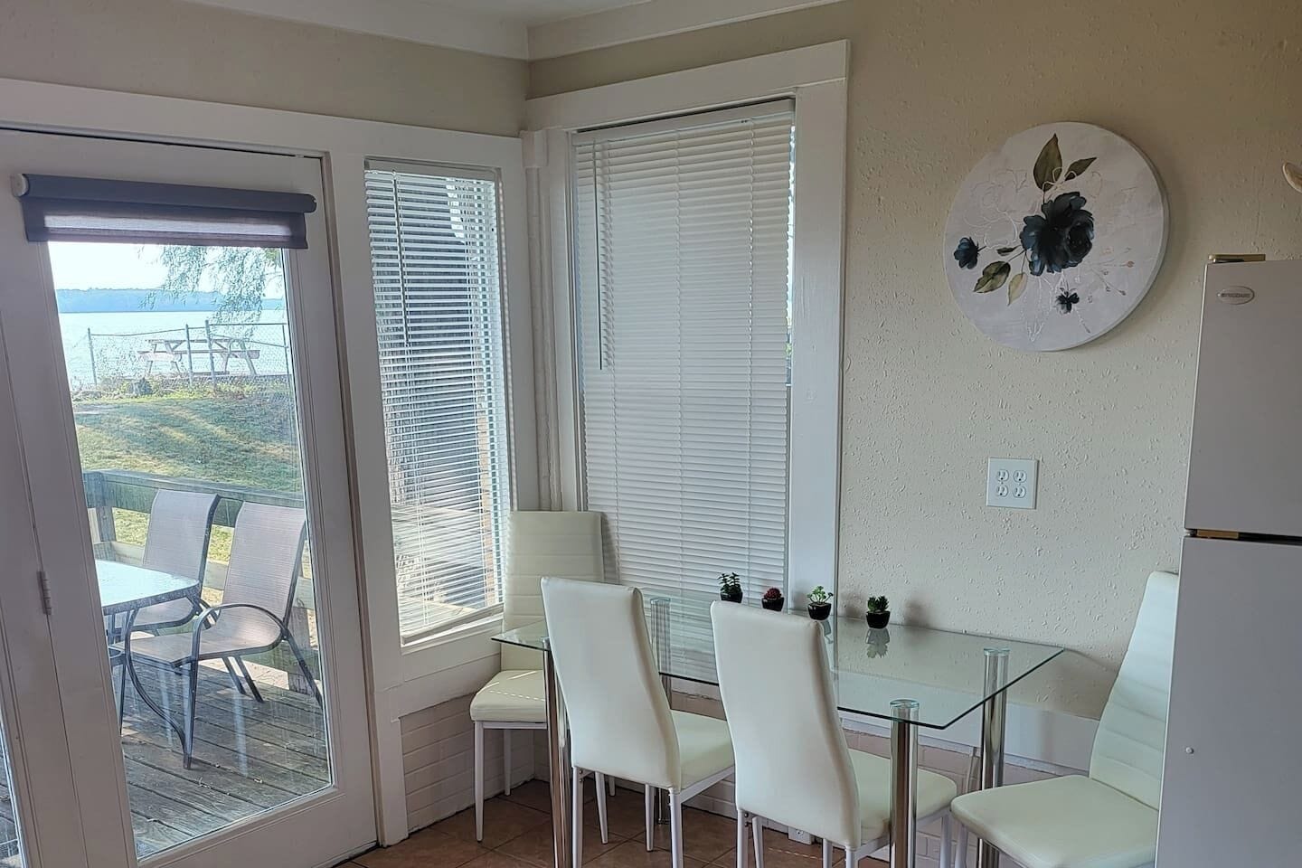 A room with a glass table and white chairs, a door leading outside, and a wall-mounted decorative plate.