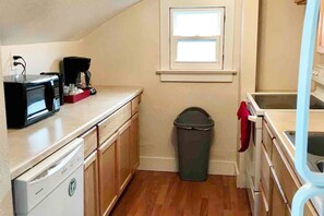 A compact kitchen with a dishwasher, microwave, and coffee maker on the counter, a trash bin, and a window above the sink.