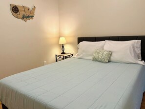 A neatly made bed with a light blue bedspread and white pillows, a small bedside table with a lamp, and a wall decoration of a wooden map with the word 'Explore'.