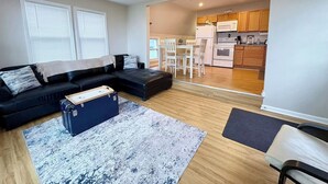 A living room with a black sectional sofa, a blue ottoman, and a patterned rug.