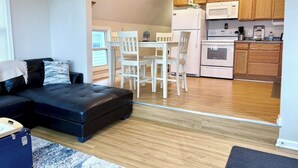 A modern living room with a black sofa, a dining table with chairs, and a kitchen area with wooden cabinets and appliances.