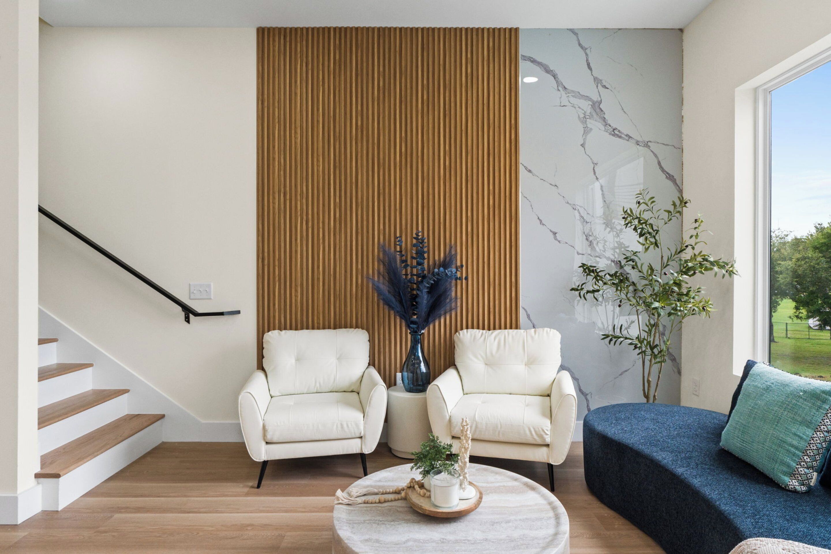 A modern living room with a staircase, two white armchairs, a blue sofa, a marble wall, and a wooden panel.