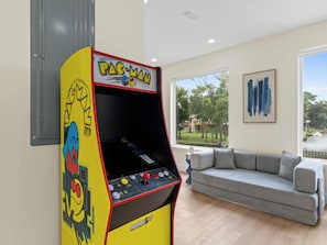 A Pac-Man arcade machine in a living room with a gray couch and a view of a lake. - 3002 Blue Lagoon C | Luxe Group Getaway, Steps to Beach, Lagoon Views (Panama City Beach)