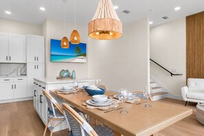 Una cocina moderna con armarios blancos, una mesa de comedor de madera para cuatro personas y un cuadro grande con una escena de playa.