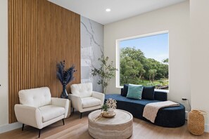 A modern living room with a sectional sofa, two armchairs, a round coffee table, and a large window offering a view of trees. - 3002 Blue Lagoon B | Steps to Beach, Lagoon Views + Community Pool (Panama City Beach)