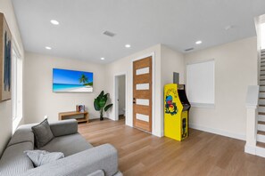 Un salón con sofá, un cuadro de una playa, una planta y una máquina recreativa amarilla.