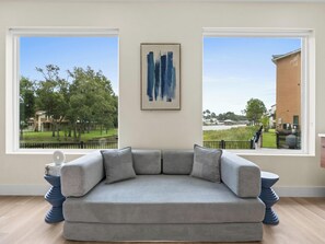 Una habitación con un sofá seccional gris, un cuadro abstracto enmarcado y ventanas amplias con vistas a un parque y edificios.