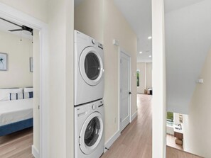 Un cuarto de lavandería con dos lavadoras apiladas, una cama con ropa de cama azul y un pasillo que conduce a un baño.