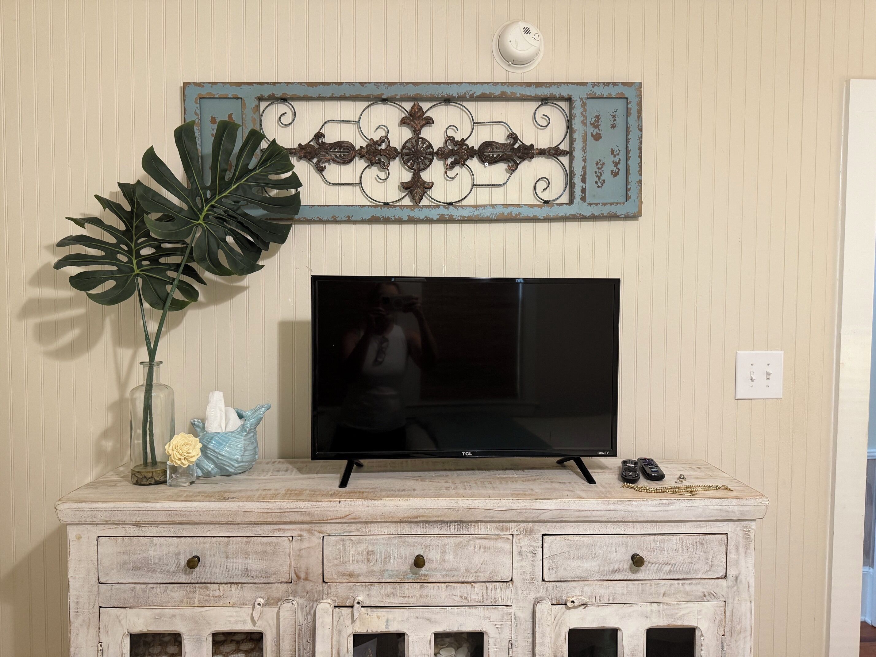 Une télévision posée sur une commode blanche avec des tiroirs, un élément décoratif en métal au-dessus, et une grande plante sur la gauche.