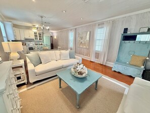 Un salon avec un canapé blanc, une table basse agrémentée d’objets décoratifs, un coin repas avec une table bleue, et une cuisine en arrière-plan.