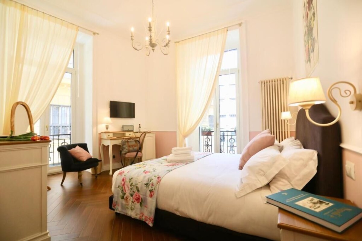 Quarto de hotel com cama, escrivaninha, cadeira, televisão, luminária e janela com cortinas.