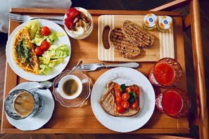 Uma bandeja de madeira com diversas opções de pequeno-almoço, incluindo torradas, uma salada, um omelete enrolado e dois copos de bebida vermelha.