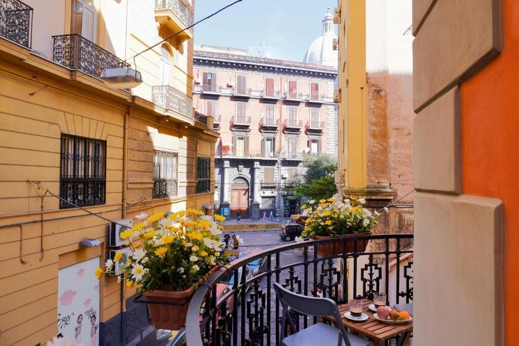 A narrow street with balconies, potted flowers, and a table with food and drinks.