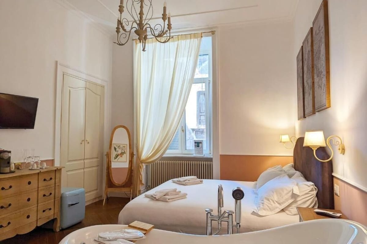 Quarto de hotel com duas camas, uma janela grande com cortinas, um candelabro, uma televisão, uma cômoda e um banheiro com banheira.