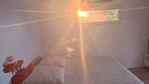 Una habitació amb un llit, una taula de fusta amb un ram de flors, i una finestra per on entra la llum del sol.