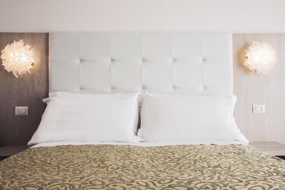 A hotel room with a tufted headboard, two white pillows, a patterned bedspread, and wall-mounted light fixtures.