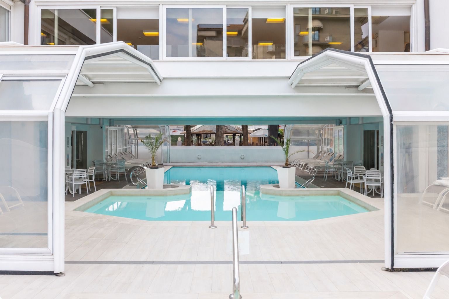 Una piscina interior con paredes de vidrio transparente, rodeada de sillas y mesas blancas, y vistas al interior del edificio.