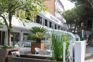A hotel exterior with outdoor seating, a palm tree, and a white fence. - Hotel Bassetti (Cervia)