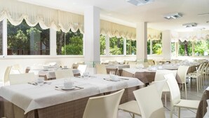 A dining area with multiple tables and chairs, large windows with curtains, and a view of greenery outside. - Hotel Bassetti (Cervia)