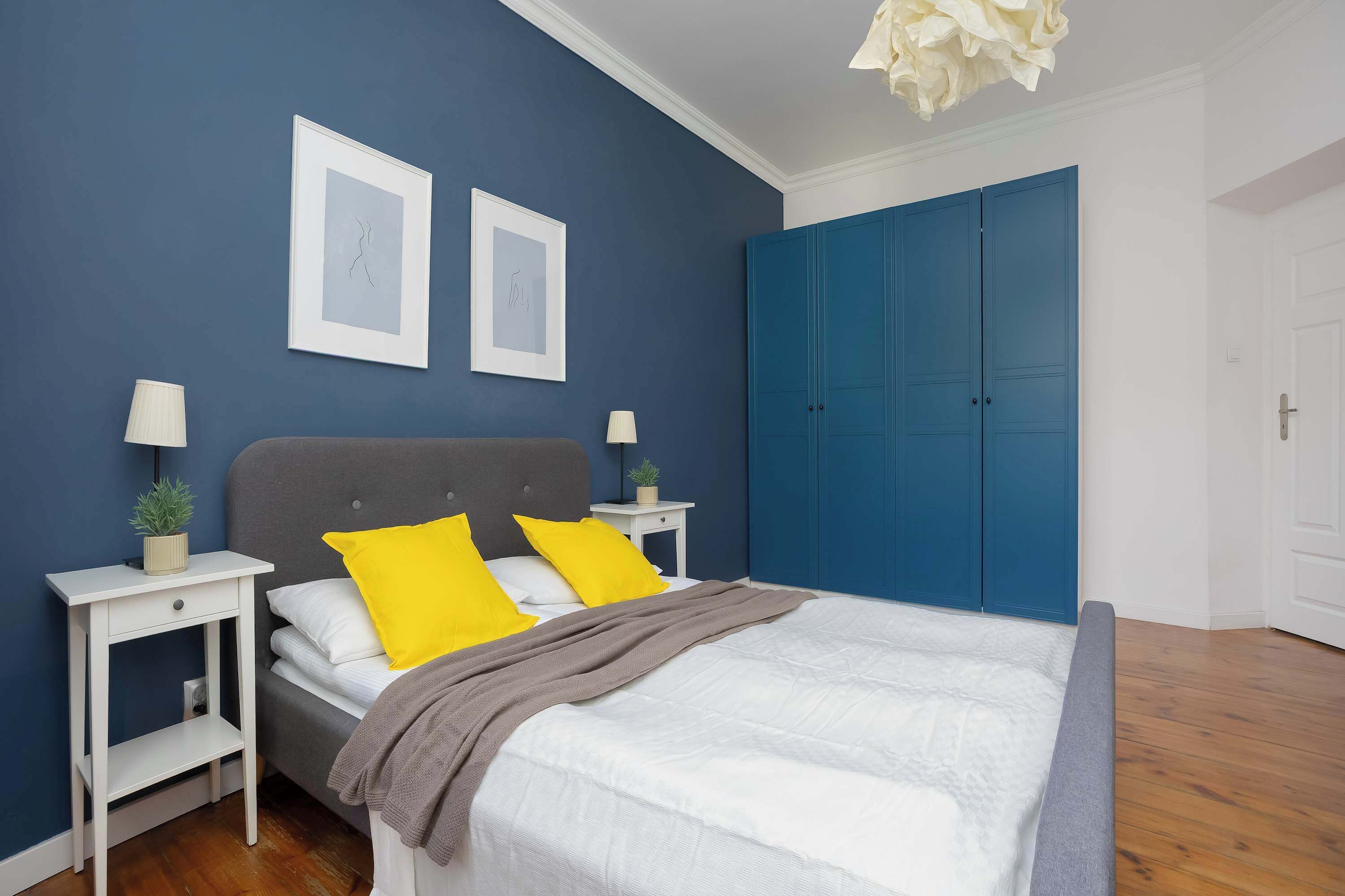 Un dormitorio con una cama, dos mesitas de noche, un armario azul y dos cuadros en la pared.
