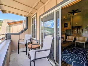 Ein Balkon mit Stühlen und einem Tisch, eine Glasschiebetür zu einem Wohnbereich mit Deckenventilator und Blick auf ein Haus im Hintergrund.