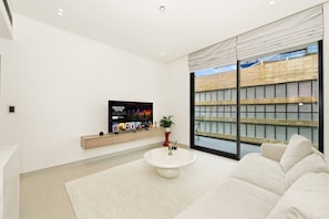 Ein modernes Wohnzimmer mit Flachbildfernseher, einer Couch, einem Couchtisch und einem großen Fenster mit Blick auf ein Gebäude.