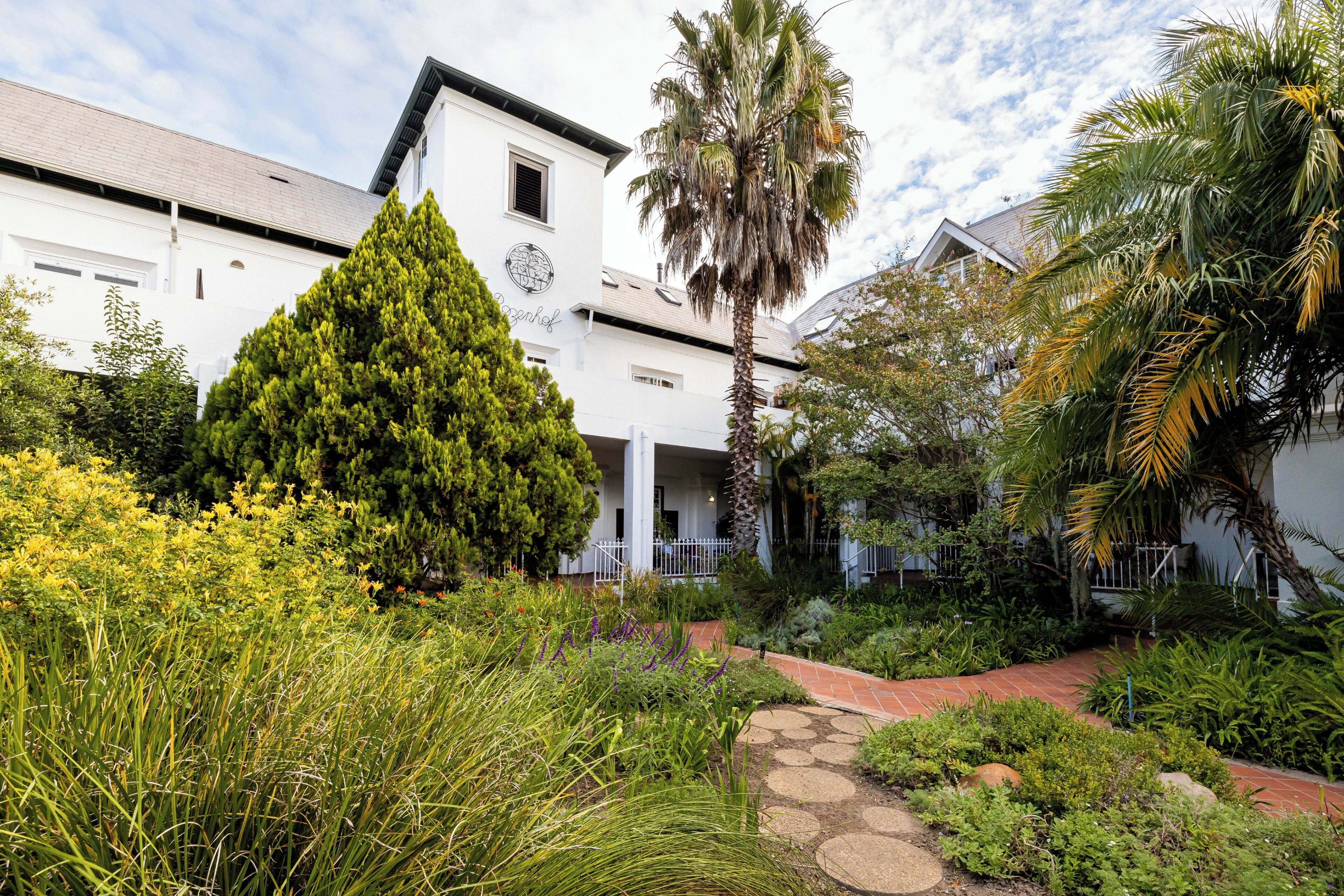 A white building with a garden in front, including a palm tree and various plants.