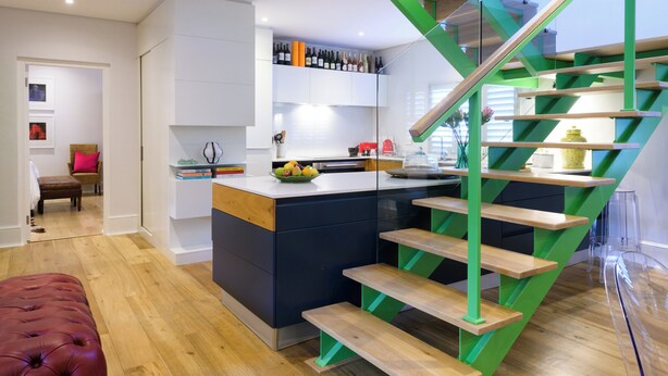 Una cocina moderna con una isla central, una escalera con detalles verdes y una zona de comedor con un banco tapizado en rojo.