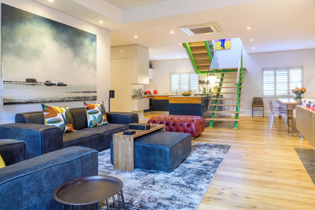 Ein modernes Wohnzimmer mit einer Couch, einem Hocker und einem Couchtisch, einem Bild an der Wand und einer Treppe mit einer gr ünen Geländer.