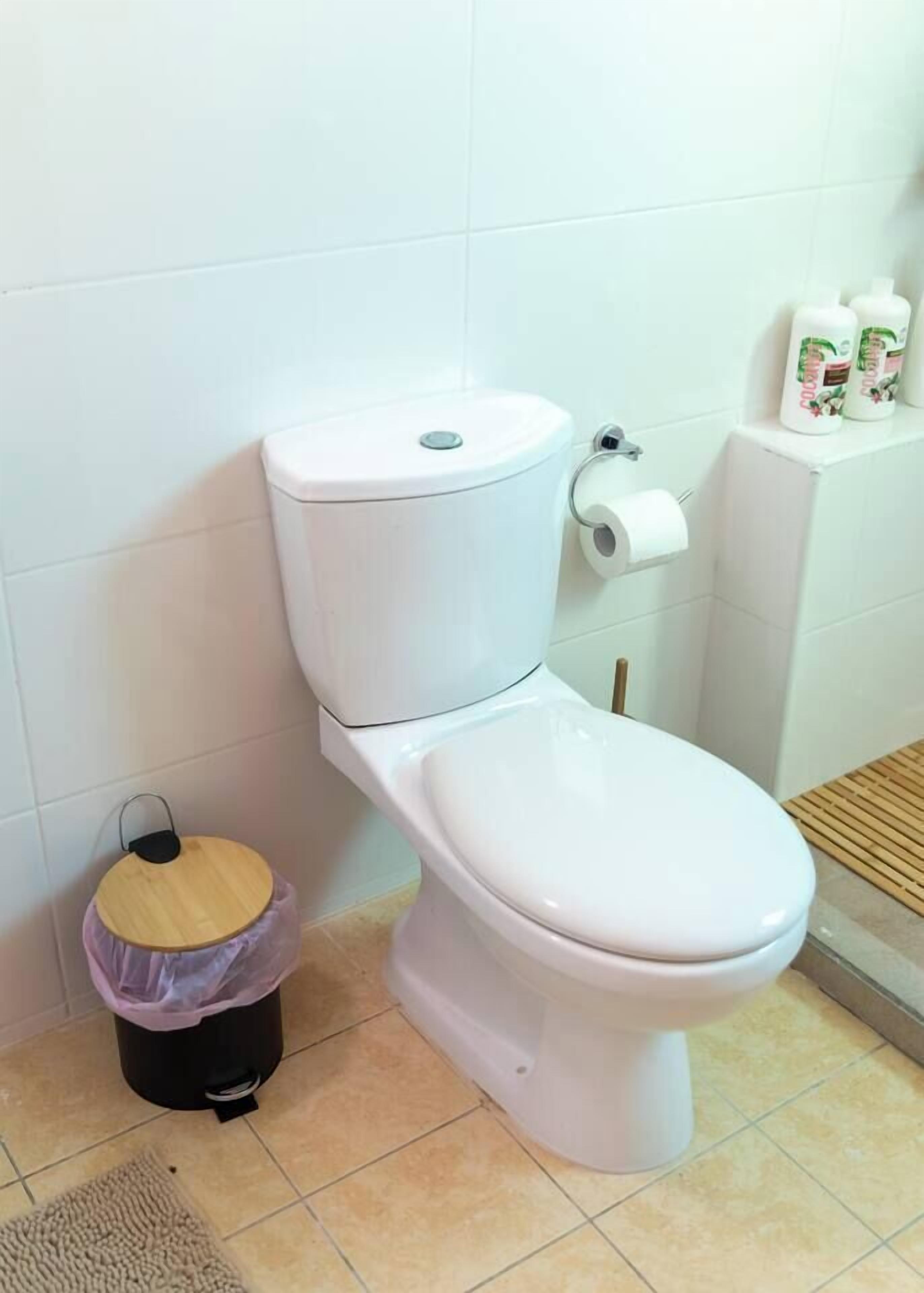 Des toilettes blanches avec un abattant, un rouleau de papier toilette et une poubelle avec un couvercle.