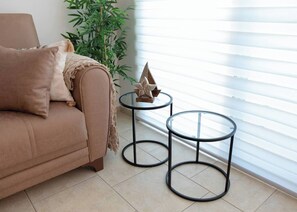 Un canapé beige avec un coussin, deux petites tables rondes à structure métallique, une étoile décorative et une plante verte dans une pièce dont le sol est recouvert de carreaux.