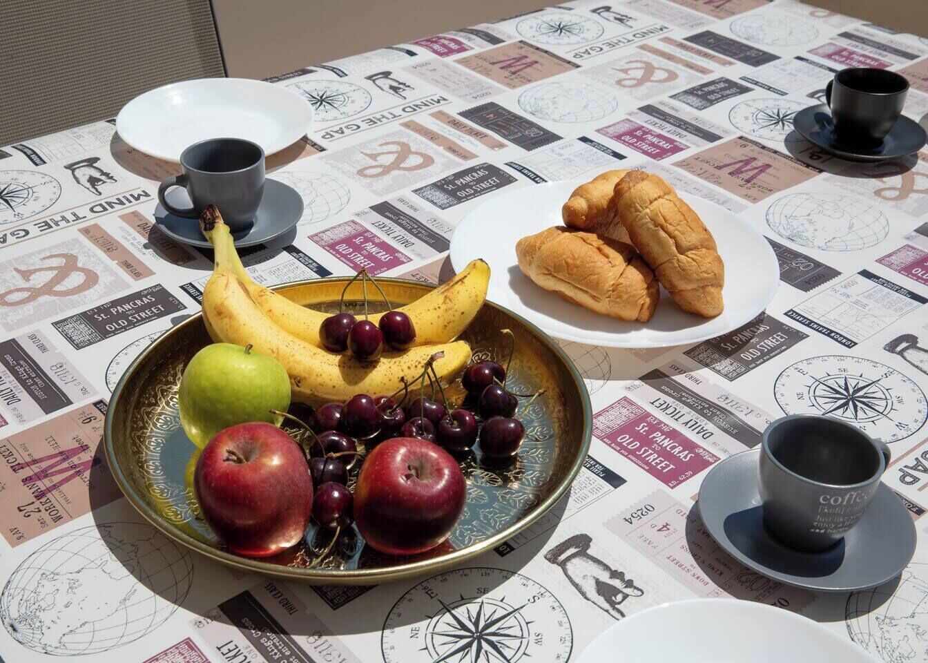 Ein Tisch mit einer Fruchtschale, einem Croissant und zwei Tassen, gedeckt auf einem Tisch mit Zeitungsmotiv.