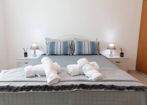 Ein ordentlich bezogenes Bett mit aufgerollten Handtüchern, einem gestreiften Kissen und zwei Nachttischen, jeweils mit einer Lampe und einem kleinen Behälter.