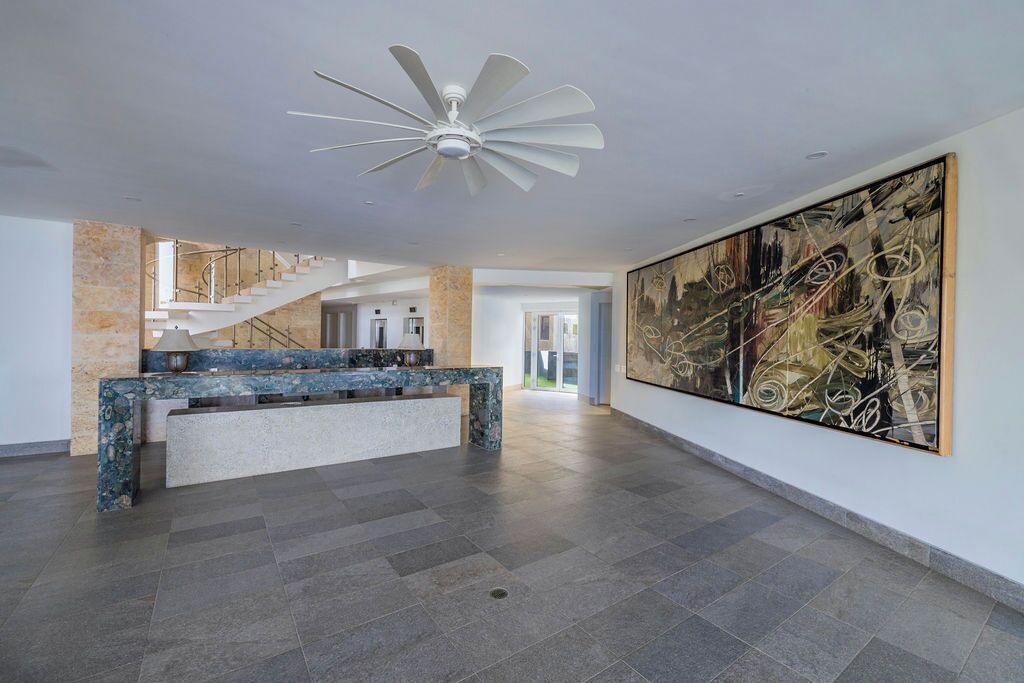 A spacious lobby with a marble reception desk, a large abstract painting, a white ceiling fan, and a staircase leading to upper floors.