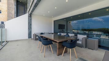 Ein moderner Außenbereich zum Essen mit einem großen Tisch und Stühlen, einer Sofagruppe und einer Glaswand mit Blick auf die Stadtlandschaft.