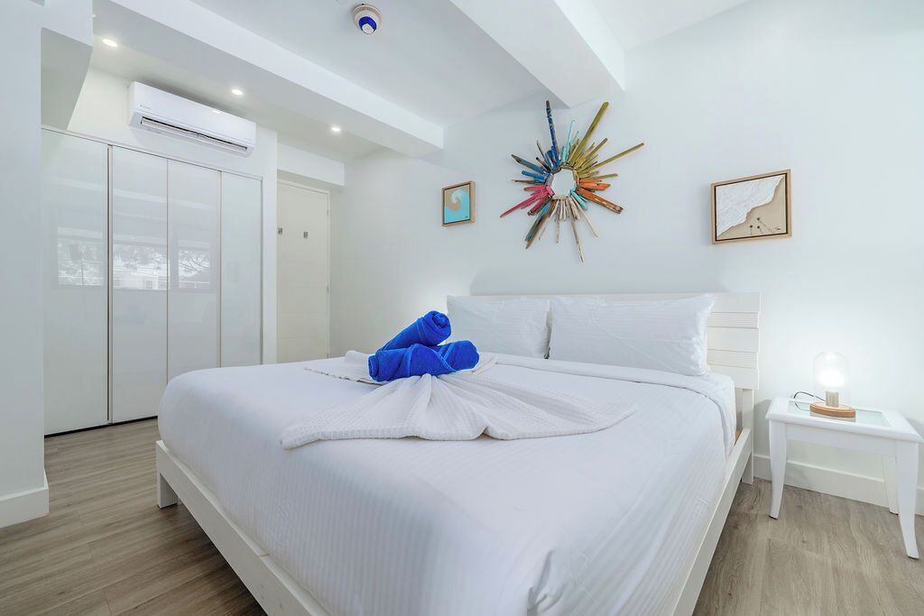 Quarto de hotel com cama, paredes brancas, ventilador de teto e um objeto decorativo na parede.