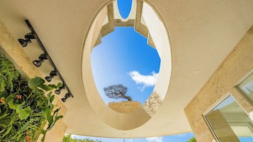 Ein architektonisches Element mit einer kreisförmigen Öffnung, umrahmt von einer Betonstruktur, durch die ein Baum sichtbar ist, sowie eine an der Wand befestigte Leuchte.