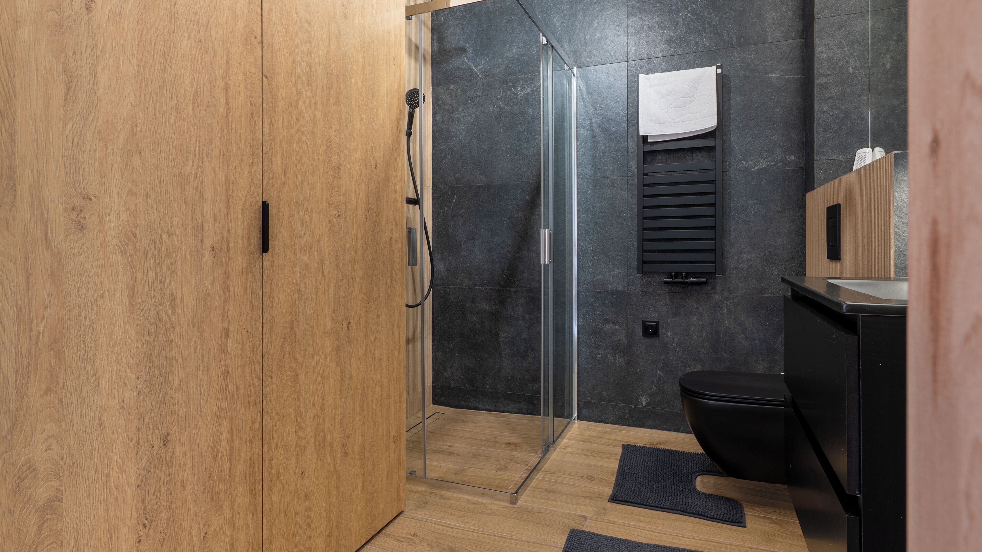 A modern bathroom with a wooden wardrobe, a glass shower enclosure, a black toilet, and a towel hanging on a rack.