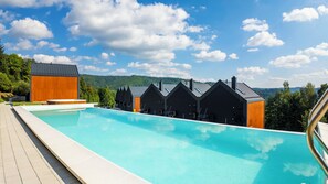 A swimming pool with a wooden deck, surrounded by black buildings with a modern design, set against a backdrop of a clear sky and green hills. - Apartamenty Sun & Snow Wisła View (Wisła)