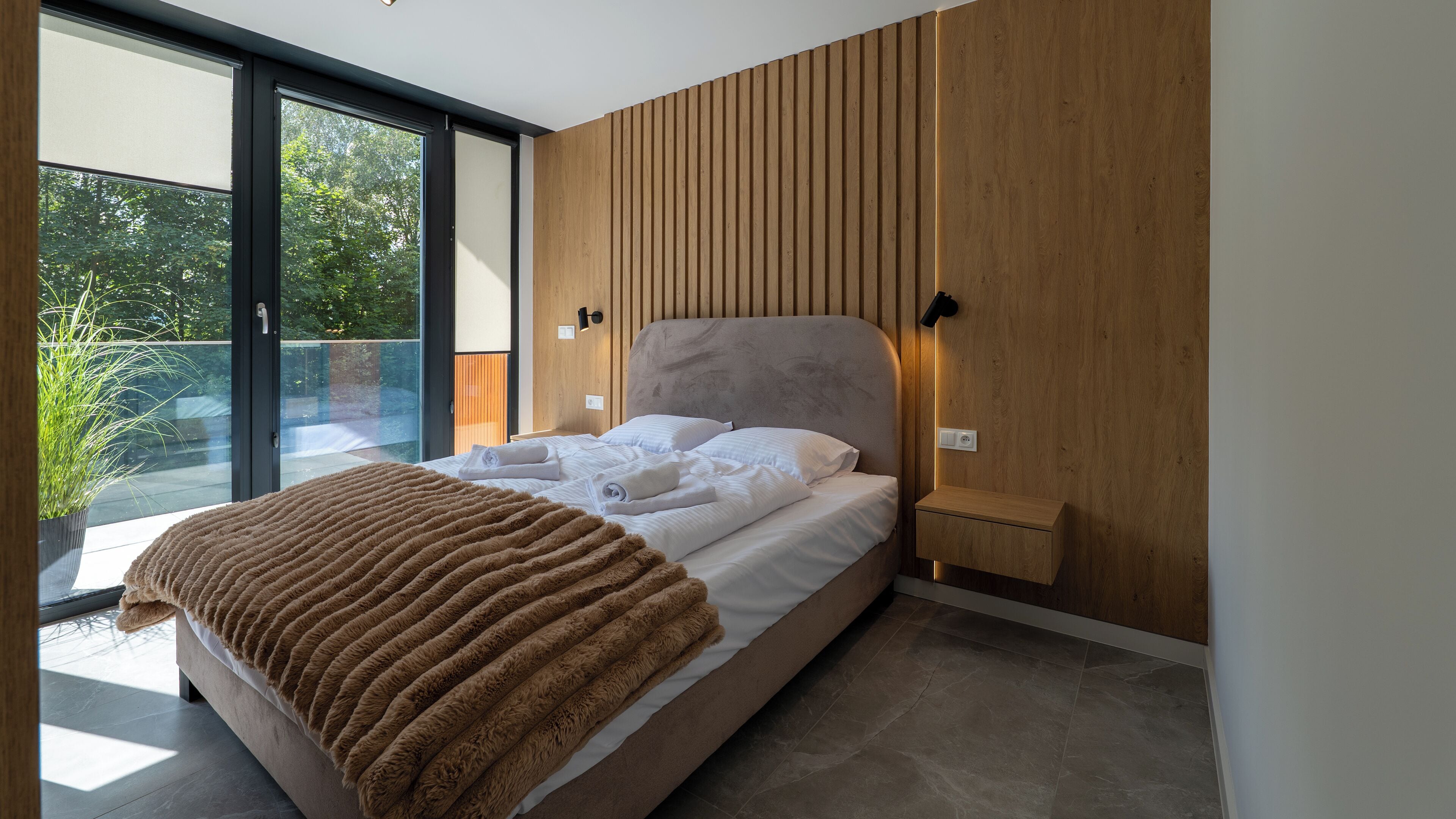 Un dormitorio moderno con una cama grande, vista a zonas verdes y una pared de madera decorativa.