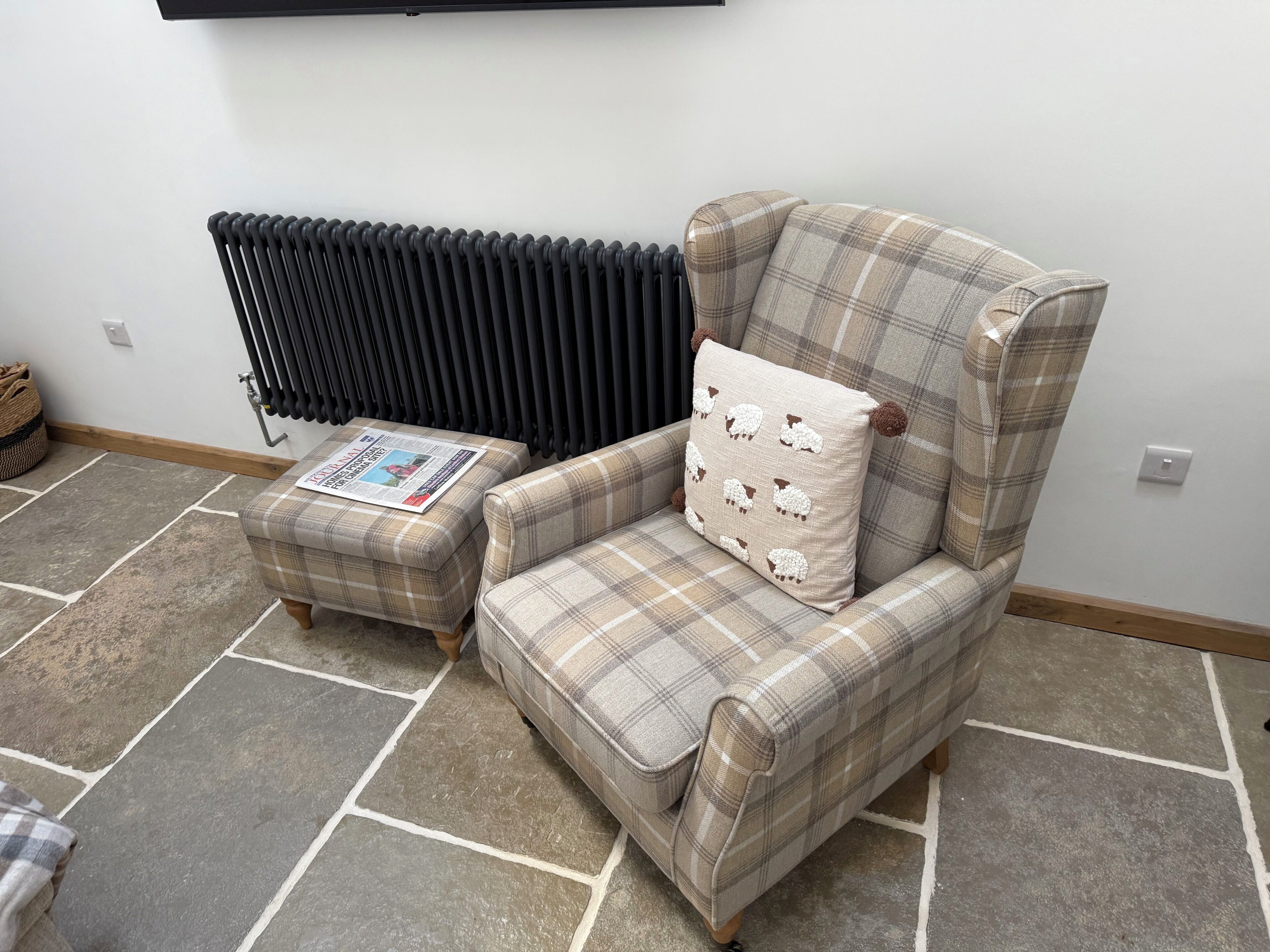 A room with a plaid armchair and ottoman, a radiator, and a newspaper on the ottoman.