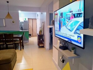 Ein Wohnzimmer mit Flachbildfernseher an der Wand, einer Essecke mit Stühlen und Tisch sowie einer Küchenzeile mit Schränken und Kühlschrank.