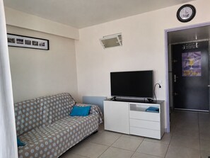 Een woonkamer met een bank, een flatscreen televisie, een klein kastje en een deur met een poster.