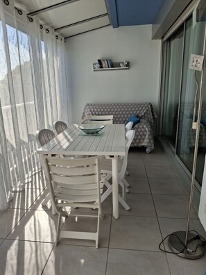 A balcony with a dining table and chairs, a sofa, and a small shelf with books and a decorative item. - Studio apartment with a loggia near the ocean (Le Grau-du-Roi)