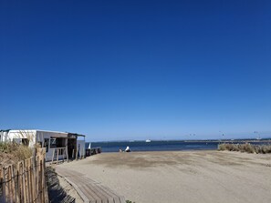 Een strand met een houten vogelzangpad, een klein wit gebouw, en een helderblauwe lucht.