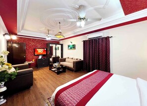 Quarto de hotel com cama, sofá, poltrona, televisão e uma mesinha com um vaso de flores.
