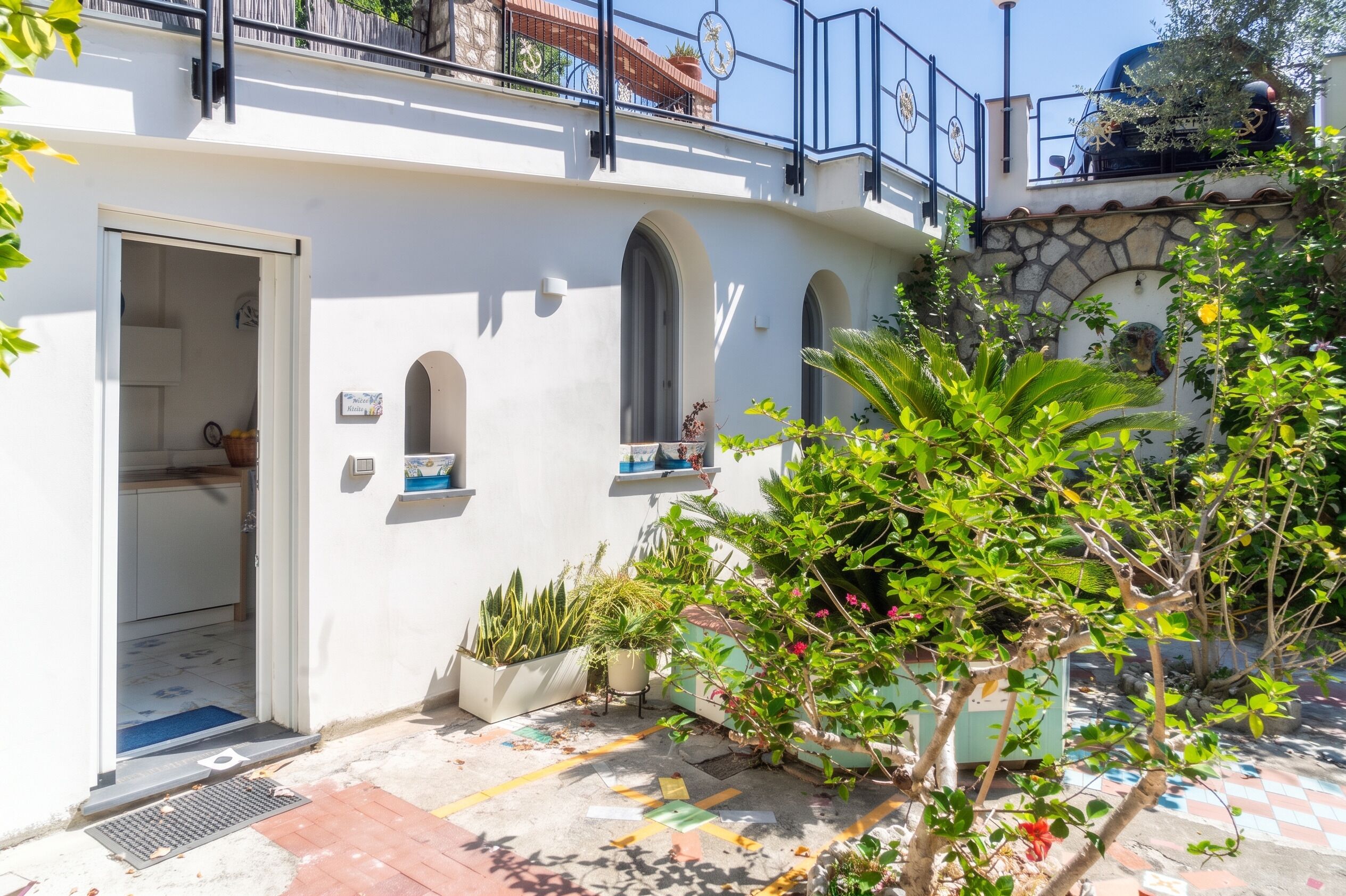 Un bâtiment à la façade blanche, doté d’un balcon avec une rambarde en métal et d’un petit jardin agrémenté de plantes en pot.