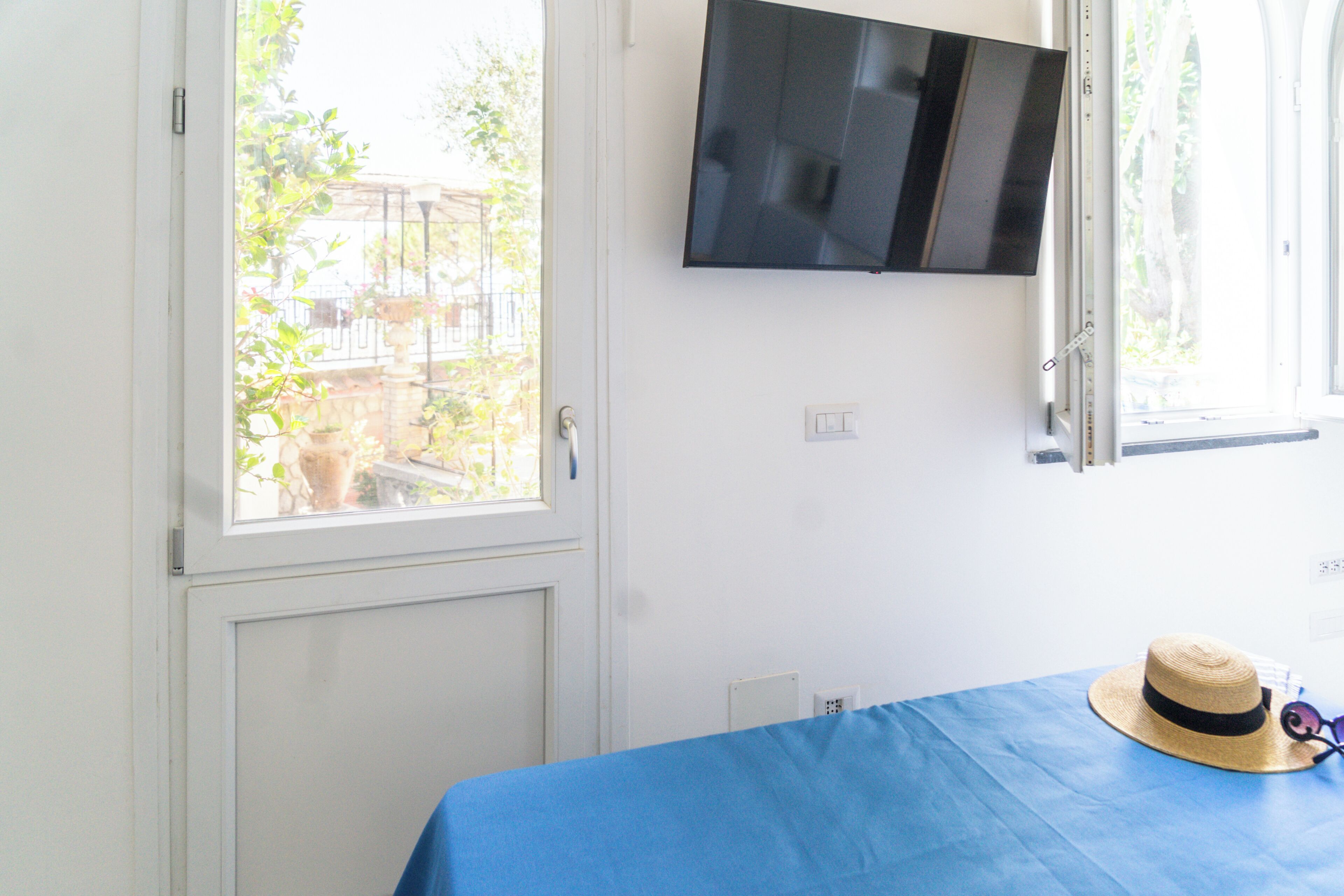 A room with a bed, a television mounted on the wall, and a window with a view of greenery outside.